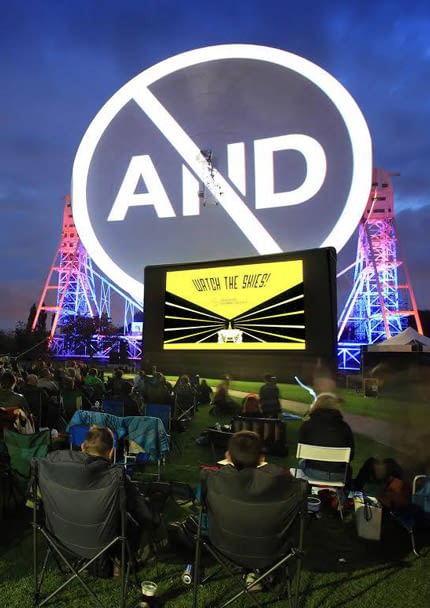 A photo of people outside sat on camping seats at Jodrell Bank, just about to catch a film at "Watch the Skies" event. The huge telescope behind the screen has the "AND Festival" logo projected on to it. The screen infront of it displays the "Watch the Skies" branding creative.