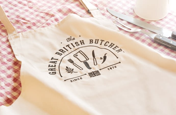 A photo of a cream apron with the Great British Butcher logo on the front, laid on a check table cloth with a carving knife and fork with wooden handles to the right.
