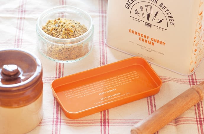 Photograph focussing on the Crunchy Curry Crumb orange tin lid. The lid is placed on a traditional looking checked tea towel, and is surrounded by the tin itself, a ceramic pot with a lid, a ramekin containing the crumb and a wooden glazing brush.