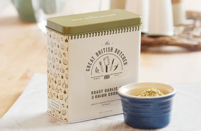 Photograph of the Roast Garlic and 3 Onion Crumb tin from the front with the lid on. The tin is rectangular shaped and has the Great British Butcher logo on the front and a pattern on the side made from repeating illustrations of the product ingredients. The tin is cream and the pattern and lid is dark green. The tin is situated in a traditional looking setting, with a dark blue ramekin containing the crumb at the front and a traditional ceramic salt and pepper pot in the background.