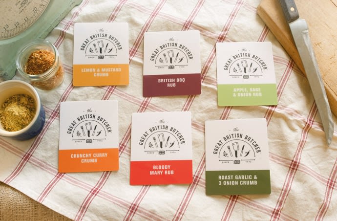Photograph of point of sale cards for use in a butchers meat counter to label meats marinated with each each flavour. The photograph is taken from above in a farm-house setting, with the cards laid out on a tea towel surrounded by pots containing the rubs / crumbs, a traditional looking weighing scale and a chunky wood chopping board and knife.