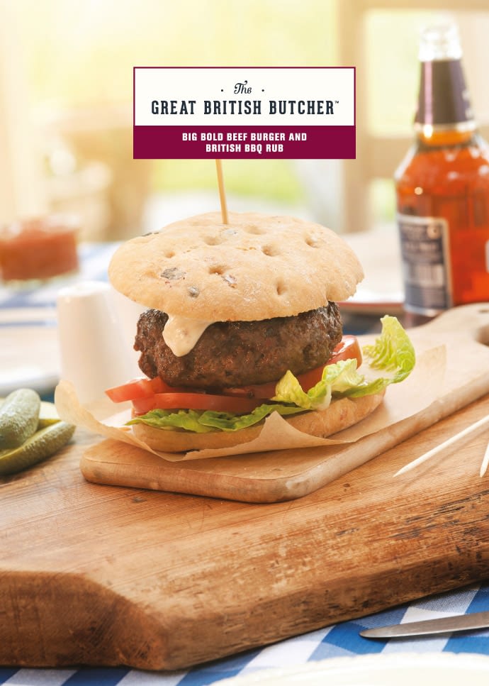 The front of a branded recipe card, presenting a chunky beef burger on a rustic bun - prepared with British BBQ Rub on a wooden service board on a farmhouse-style table with a bottle of ale in the background.
