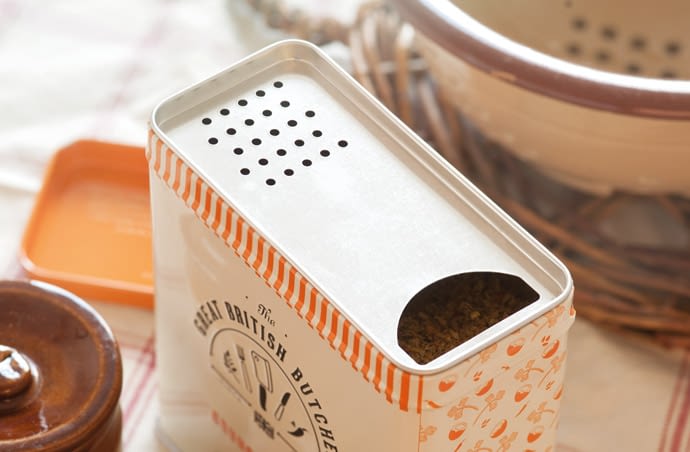 Photograph of the Crunchy Curry Crumb tin from an above angle. The tin is rectangular shaped with a pouring opening on one side and a shaker opening on the other. It has the Great British Butcher logo on the front and a pattern on the side made from repeating illustrations of the product ingredients. The tin is cream and the pattern is orange. The tin is situated in a traditional looking setting, with a ceramic colander on one side and a ceramic pot with a lid on the other. In the background is the orange tin lid.