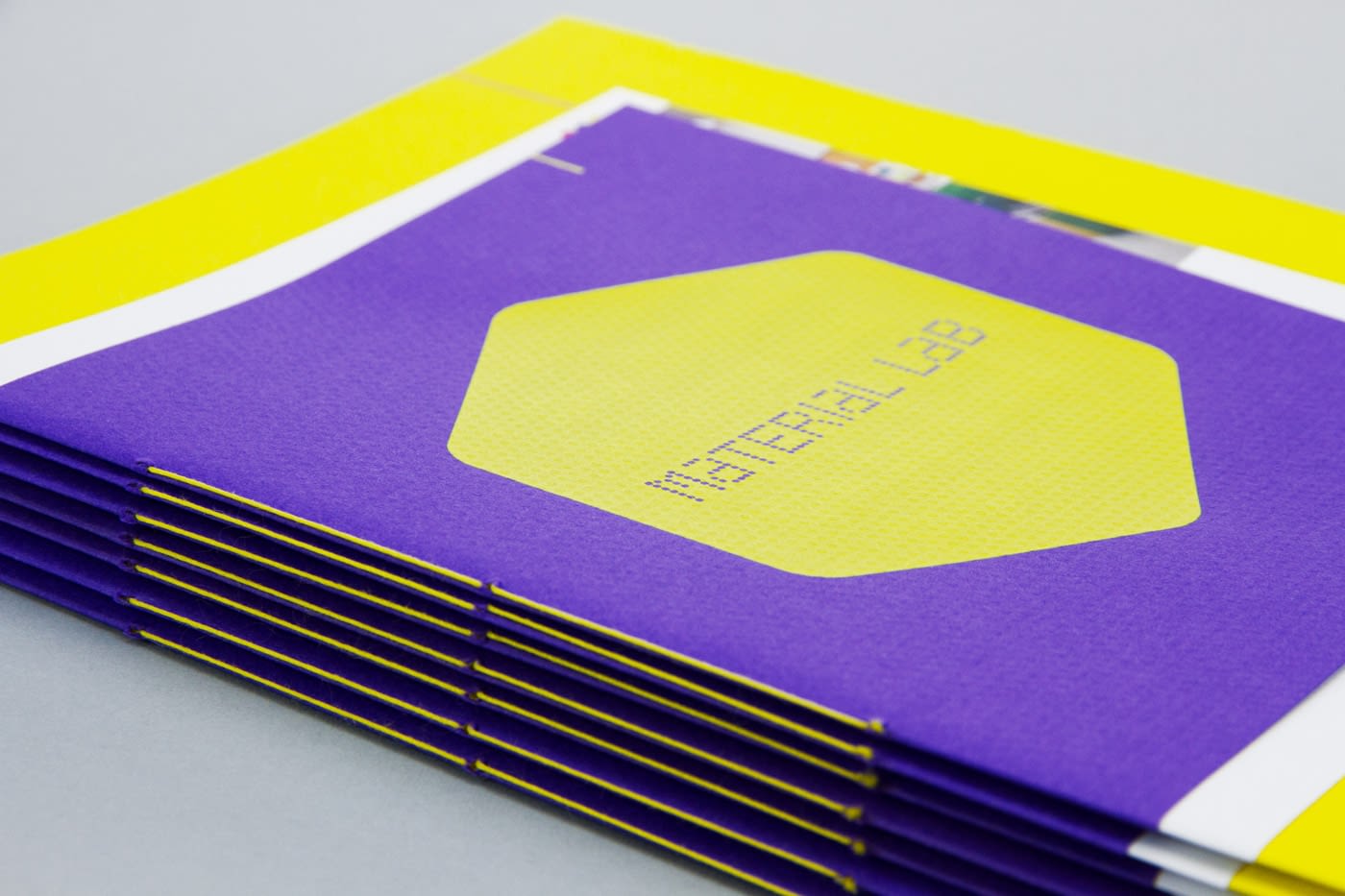 A stack of 7 Material Lab 20 page booklets. The cover stock is Colorplan Matrix emboss, in purple. On the cover is a large yellow screen printed hexagon with the Material Lab logo inverted in the centre. The sides of the booklet are visible and are saddle stitched with yellow thread.